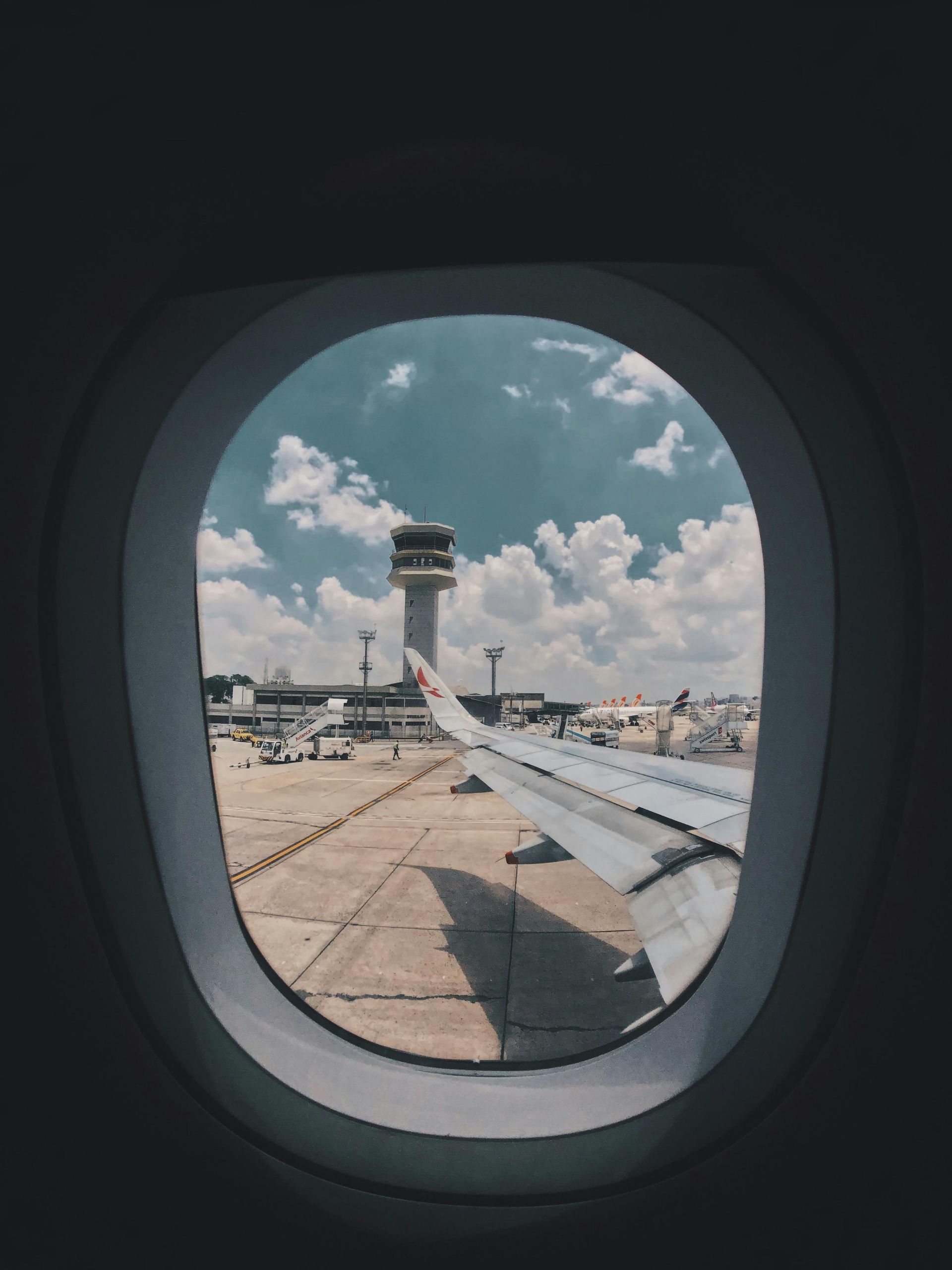 View from an airplane window: airport control tower, wing, sunny sky with clouds.