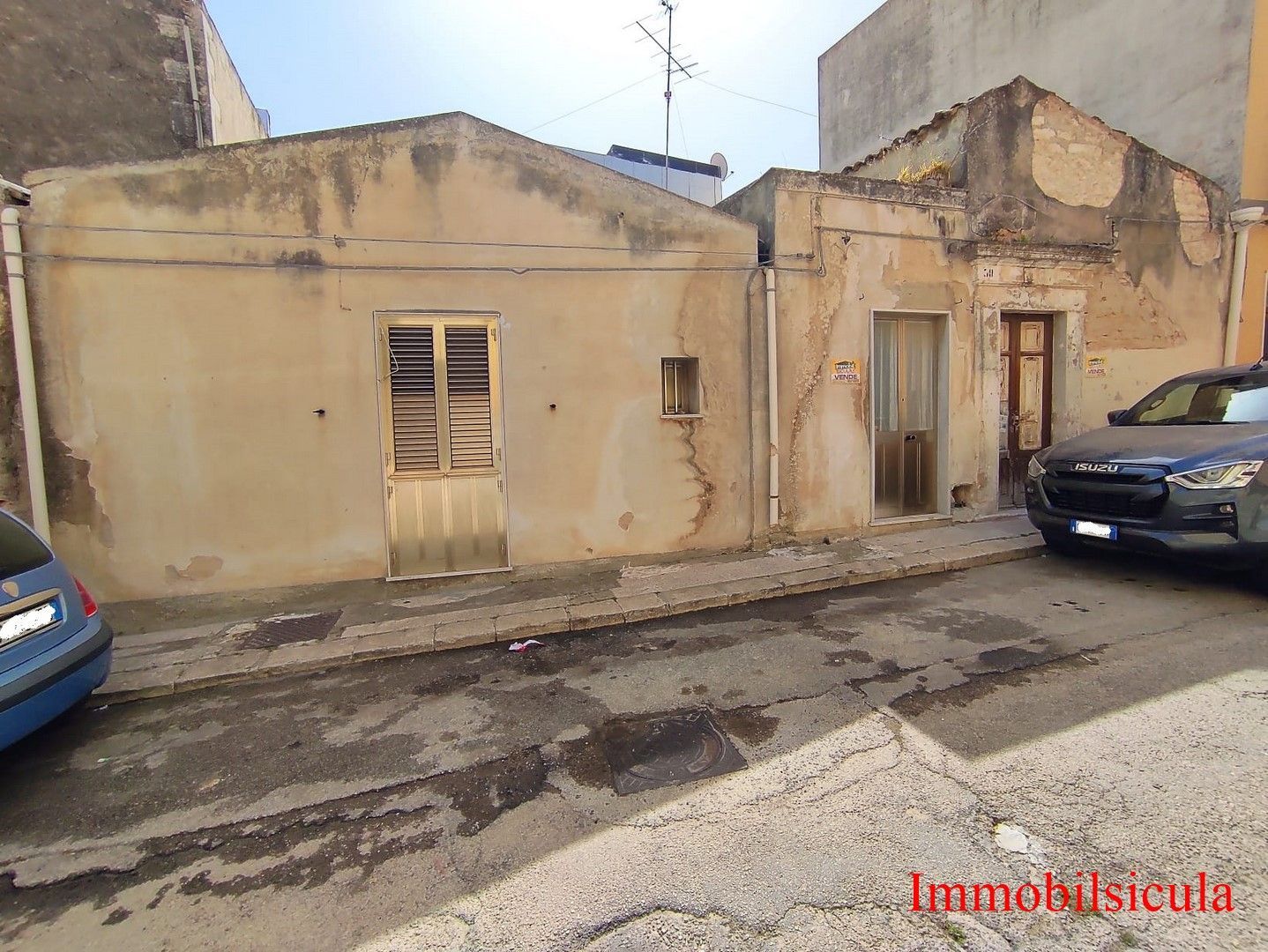 Vecchia casa beige in una strada malandata, con auto parcheggiate e muri scrostati.