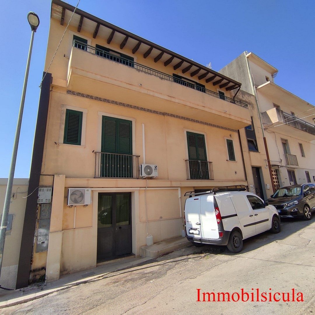 Edificio beige a tre piani con persiane verdi su una strada stretta, con un furgone bianco parcheggiato davanti.