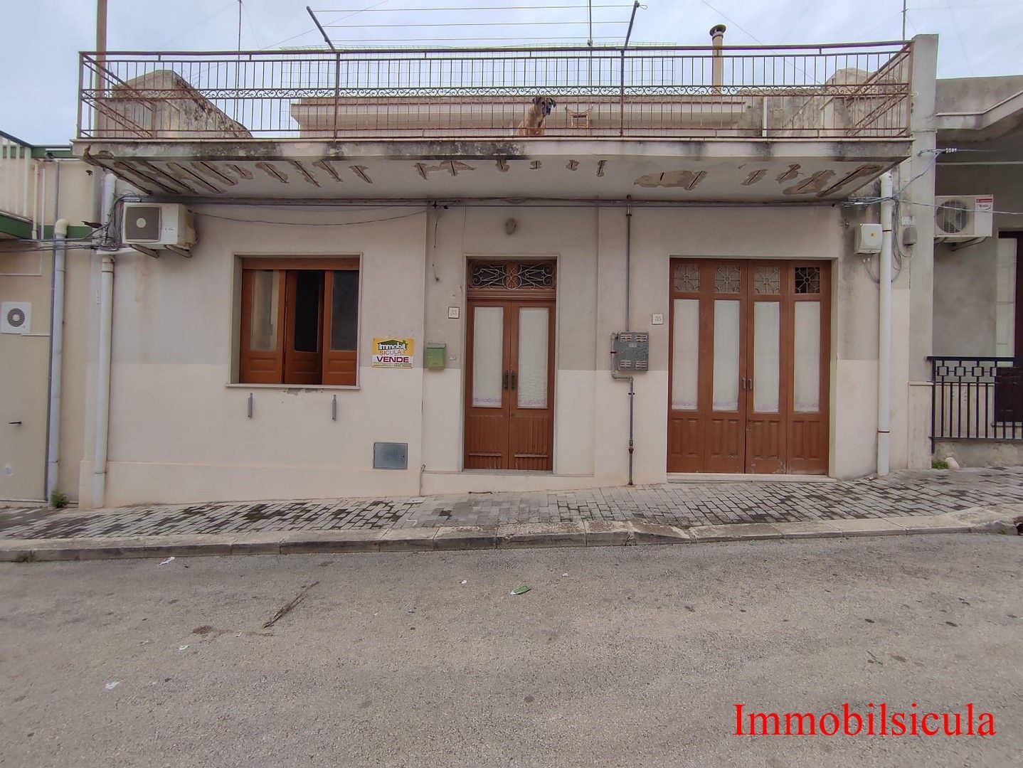 Vista frontale di una piccola casa beige con porte e finestre in legno e un balcone in metallo al piano superiore.