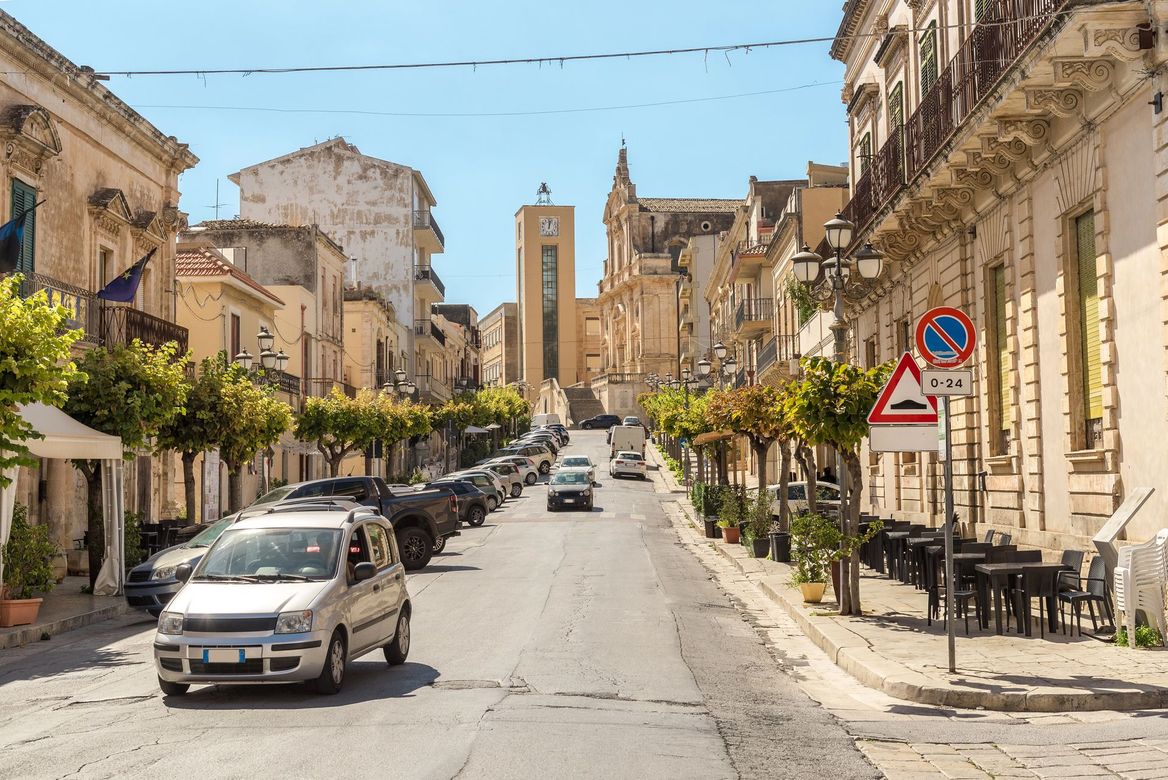 Via del centro storico illuminata dal sole, con auto, caffè ed edifici in pietra che conducono a una torre in lontananza.