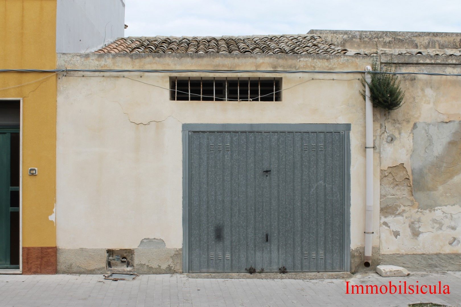 Facciata dell'edificio color beige sbiadito dagli agenti atmosferici, con un grande portone da garage grigio e una piccola finestra con le sbarre sopra di esso.