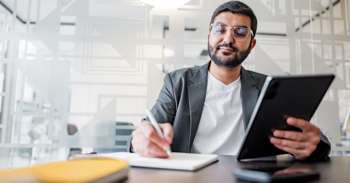 A man is sitting at a desk. He’s holding a tablet in one hand and writing something down with a pen in the other.