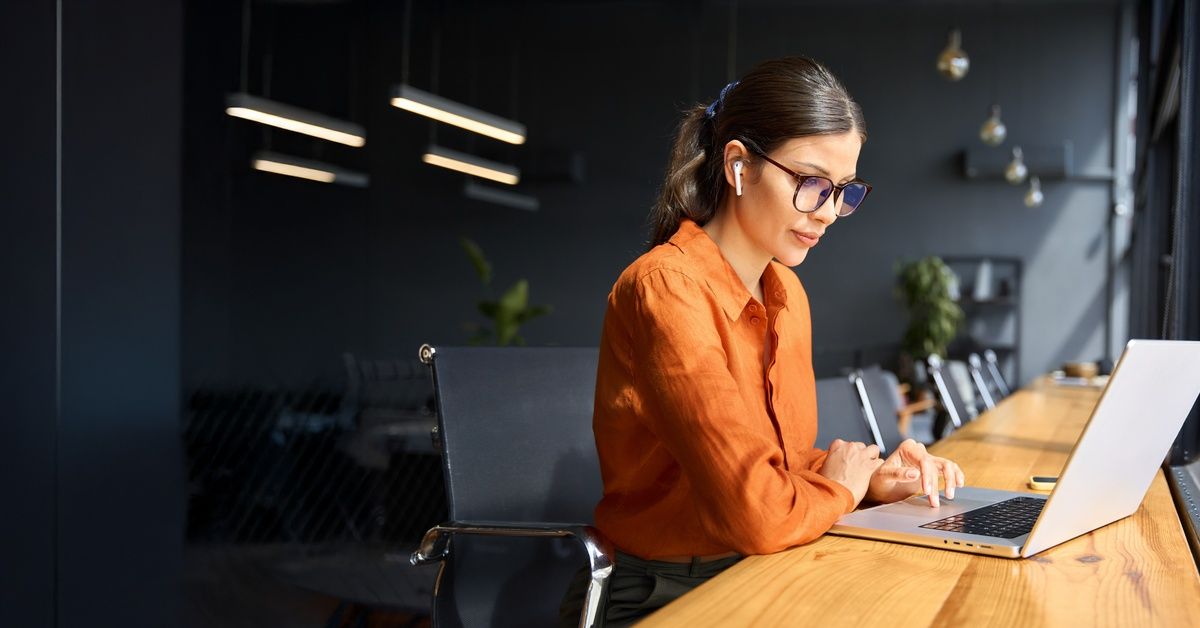 A woman with tied-back hair and professional attire is sitting at a table. She is looking at a computer screen.