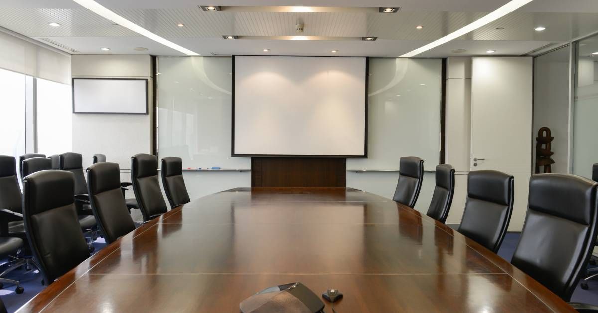 A long wooden table with several office chairs sits in front of a projector screen in an executive c