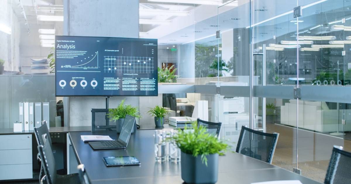 A modern office conference room furnished with a table, chairs, plants, and a large screen surrounded by glass walls.