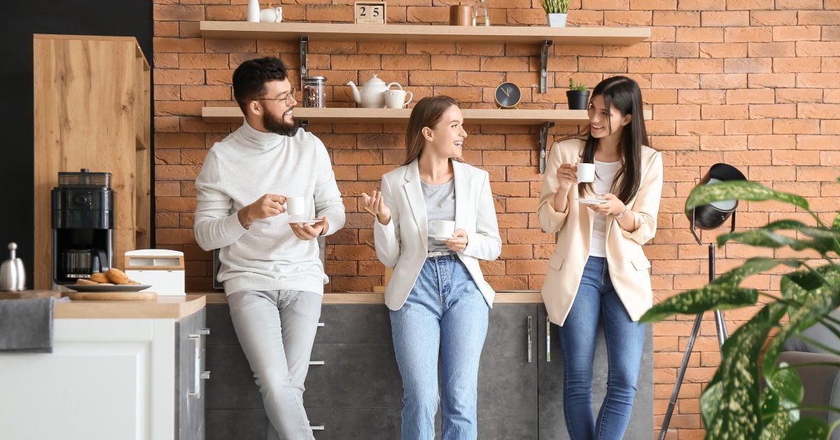 A kitchen space in an office environment with three employees standing and talking while drinking coffee.