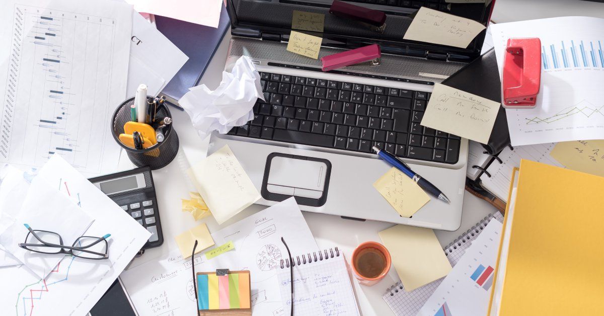 A desk with an open laptop surrounded by disarrayed sheets of paper, pens, sticky notes, and a pair of glasses.