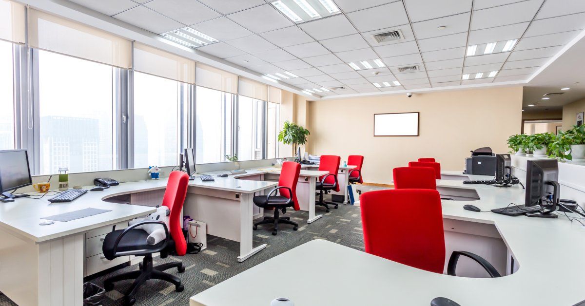 A large office space with several connected U-shaped desks with red office chairs, computers, and windows.