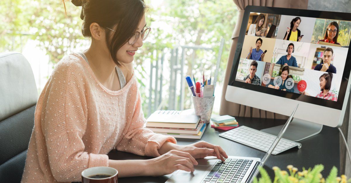 A woman sits at her desk at home and types on a laptop while taking a video conference call on a desktop computer.