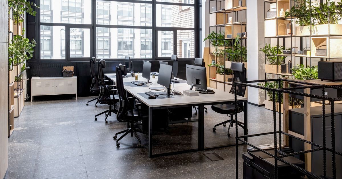 A modern loft office space with industrial features, large windows, shelves, plants, and several workspaces.