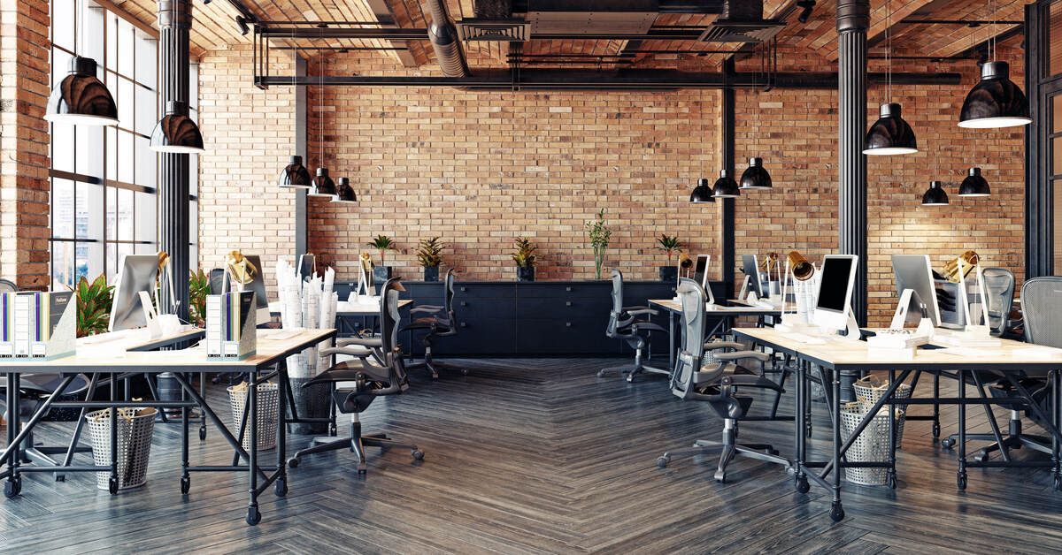 A large, open shared office space with brick walls, tall windows, several tables, swivel chairs, and