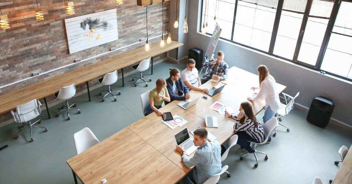 Seven people are around a large table; one of them is standing while the rest are sitting on chairs.