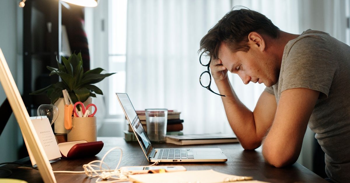 A man is sitting at a desk and is holding a pair of glasses in his hand. There is a laptop in front of him.