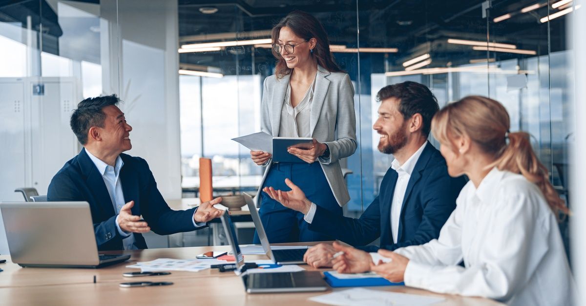 Four people are in a meeting room; three people are sitting at a table while one person is standing up.