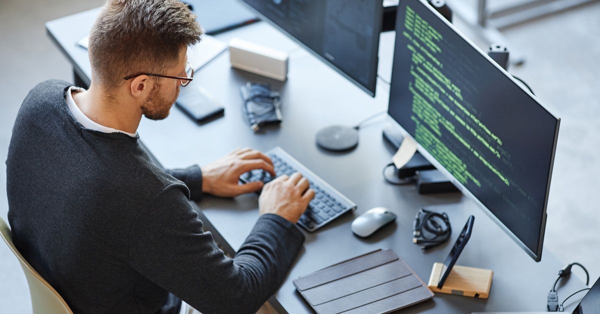 A man in a black sweater is sitting at a desk with two computer monitors. On the screens are lines of code.