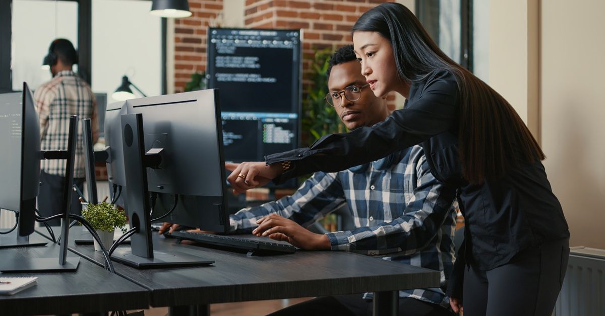 A woman is pointing at a computer monitor as a man wearing glasses sits in a desk next to her.