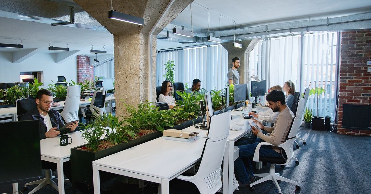 A room has several tables lined up, and on those tables are computer monitors. There are people sitting at those monitors.