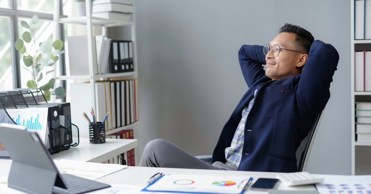 A man in an office leaning back in his chair with both hands behind his head looking out the window.