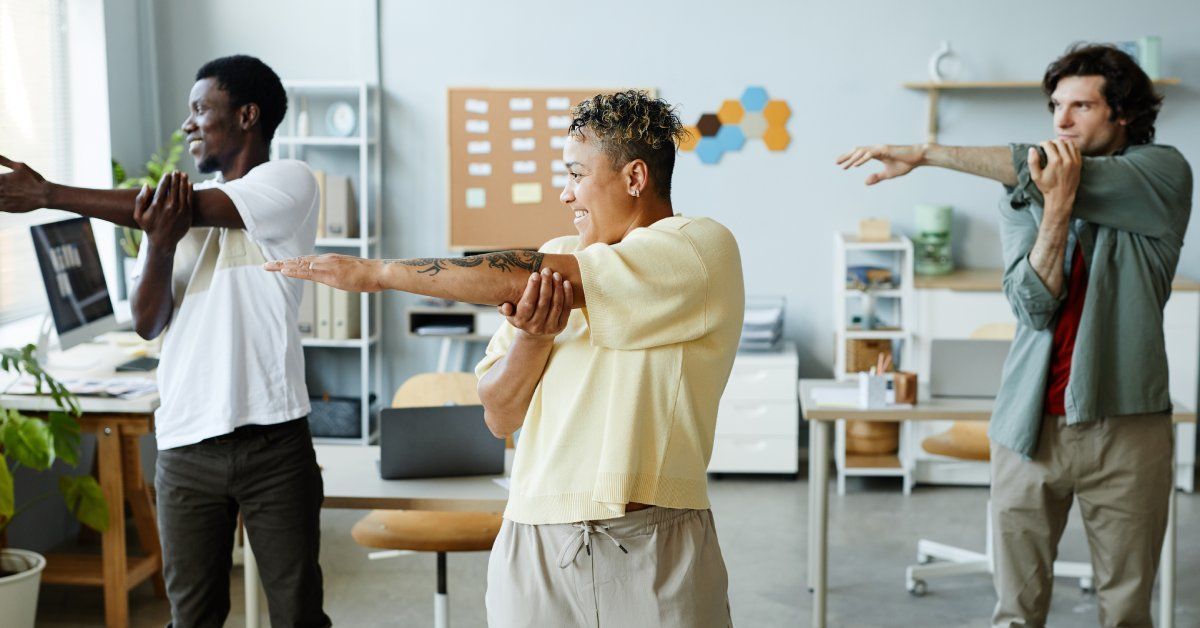 Three people in an office setting standing and holding their arms straight horizontally across their bodies in a stretch.