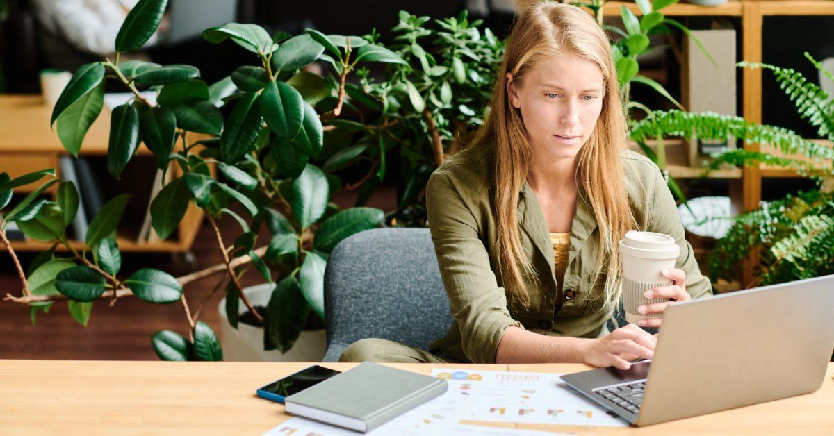 A woman sitting at a desk with an open laptop in front of her and multiple plants behind her as she holds a coffee cup.