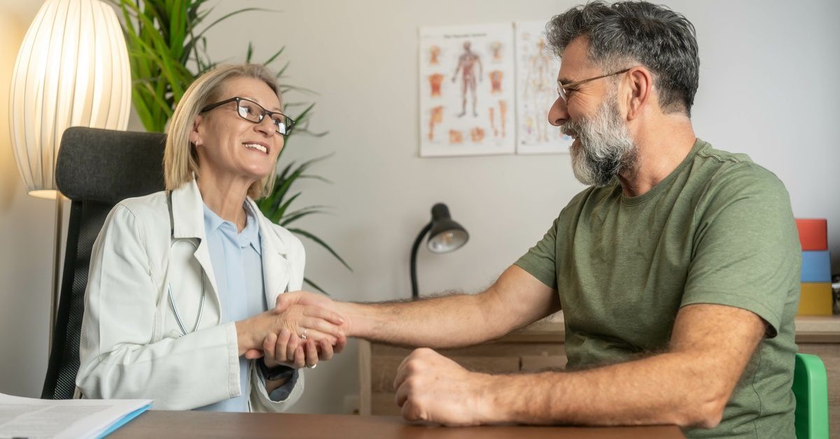 A doctor shaking hands with an older patient wearing a green shirt and glasses in an office medical posters.