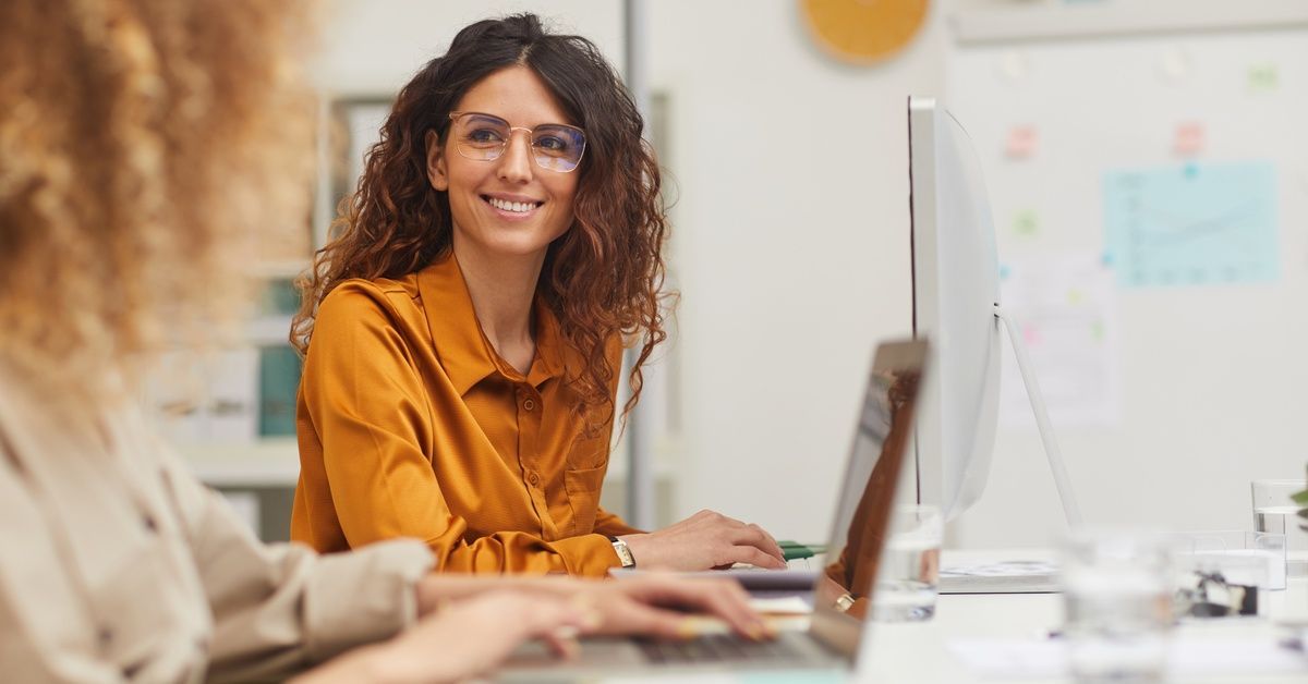 Two women facing each other in an office setting with their laptops and computers in front of them as the one in glasses smiles.