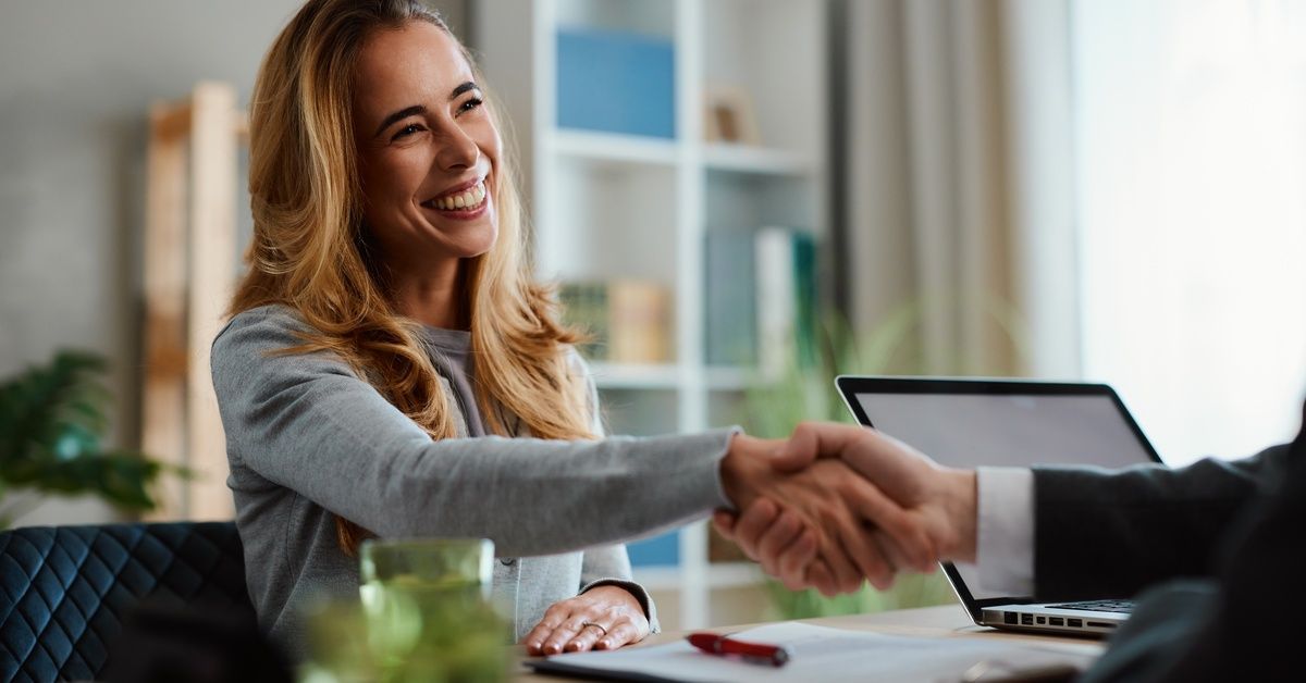 A woman smiling as she shakes the hand of someone sitting across from her at a desk.