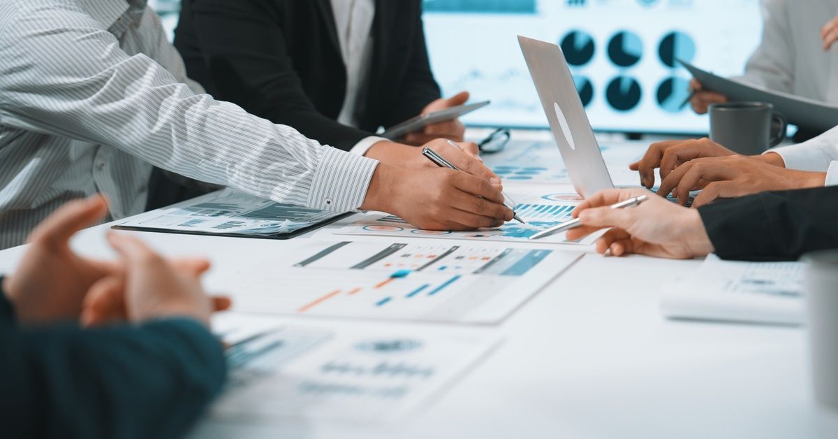 A group of people sit around a table in an office environment and point to various printed graphs and reports.