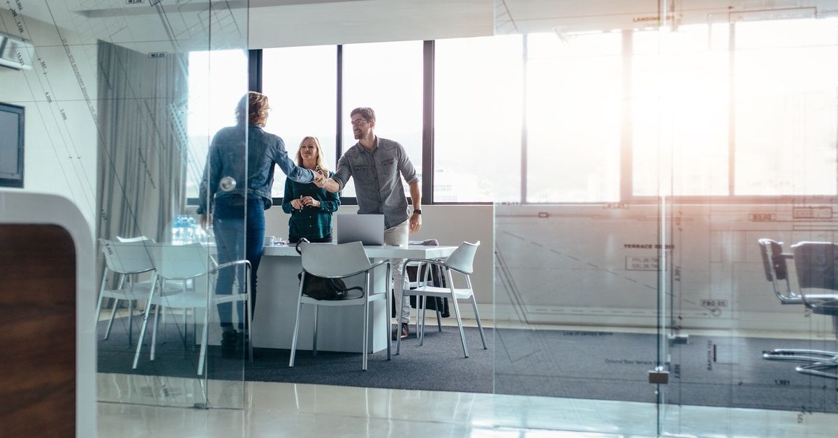 Three people gather in a small conference room with glass walls and doors surrounded by light.