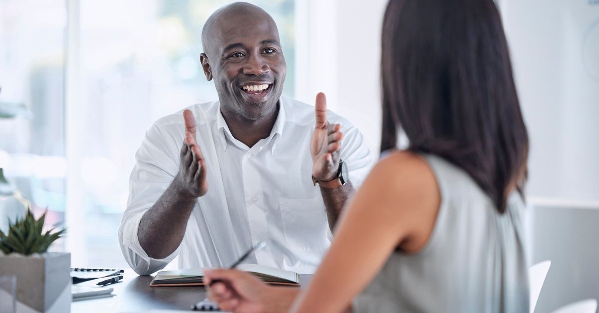 A man and woman sit across from each other and talk at a table in an office with notebooks in front of them.