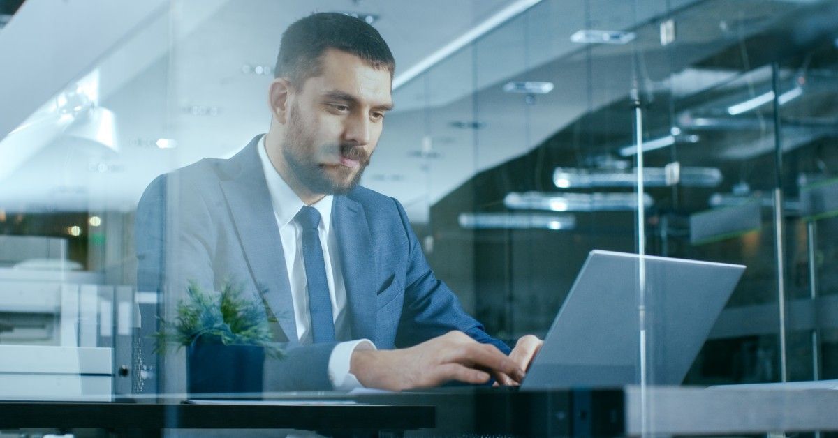 A businessman works on his laptop while facing a glass wall that reflects the rest of the office he is in.