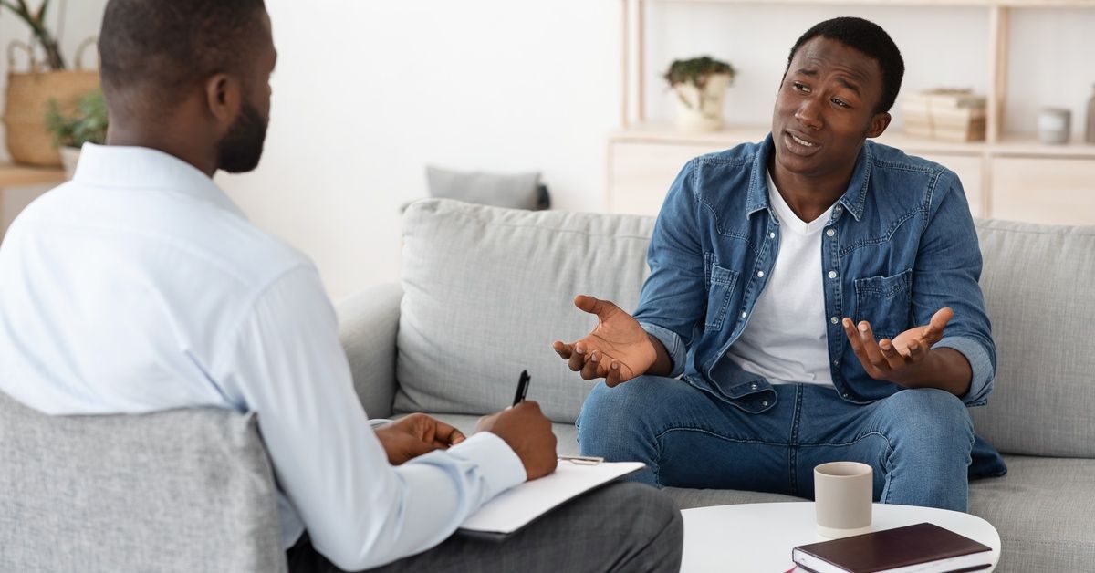 A man sitting on a couch speaks to a therapist in a room with a minimalist design and neutral colors.