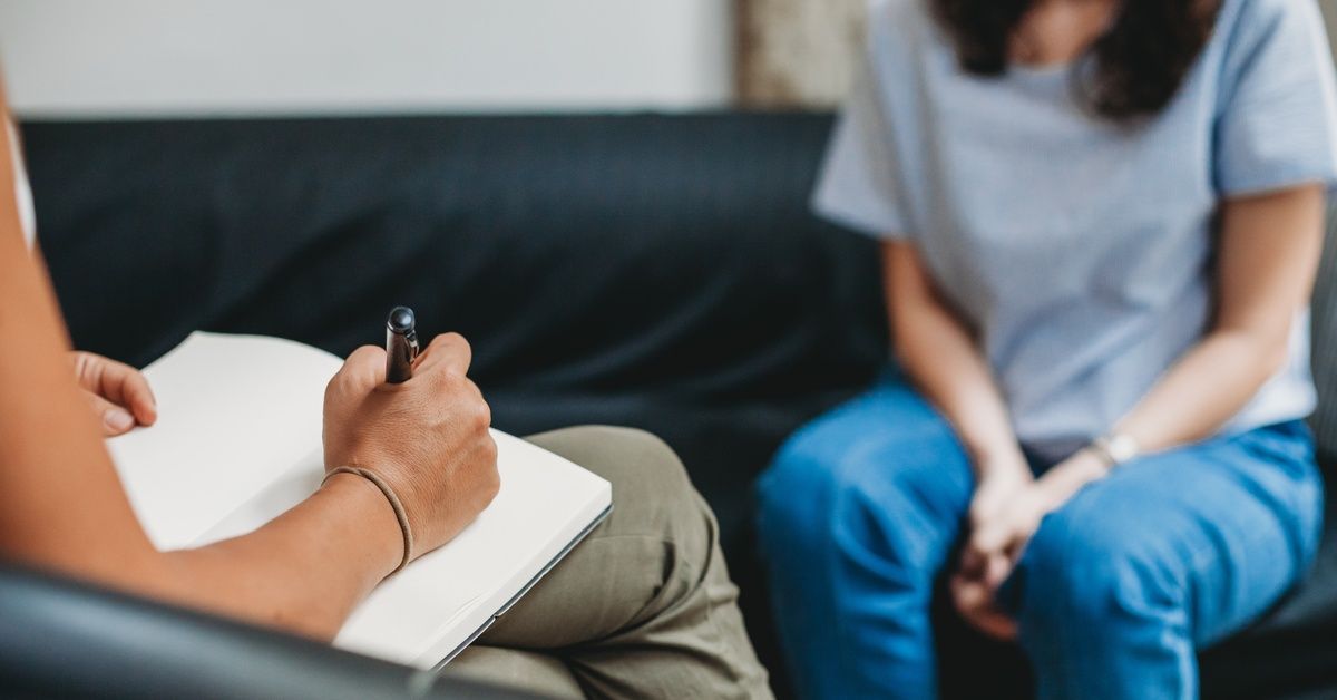 A woman sits on a couch and speaks to a therapist across from her that is jotting down notes.