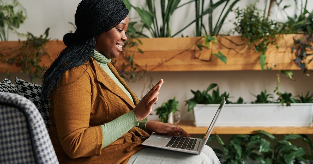 A woman sitting in a chair within an office space filled with plants holds a video call on her laptop.