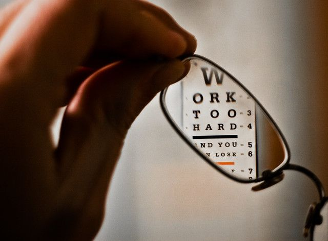 Hand holding eyeglasses, partially obscuring an eye chart with the words
