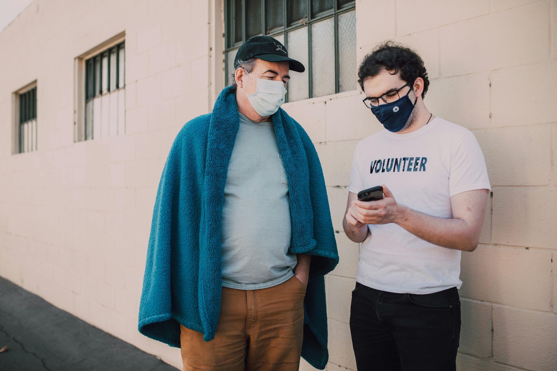 Two people wearing masks, looking at a phone outside a building. One is wrapped in a blue blanket.