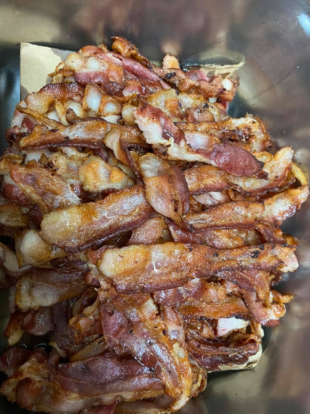 Pile of crispy, cooked bacon strips in a container; browned, with some fat.