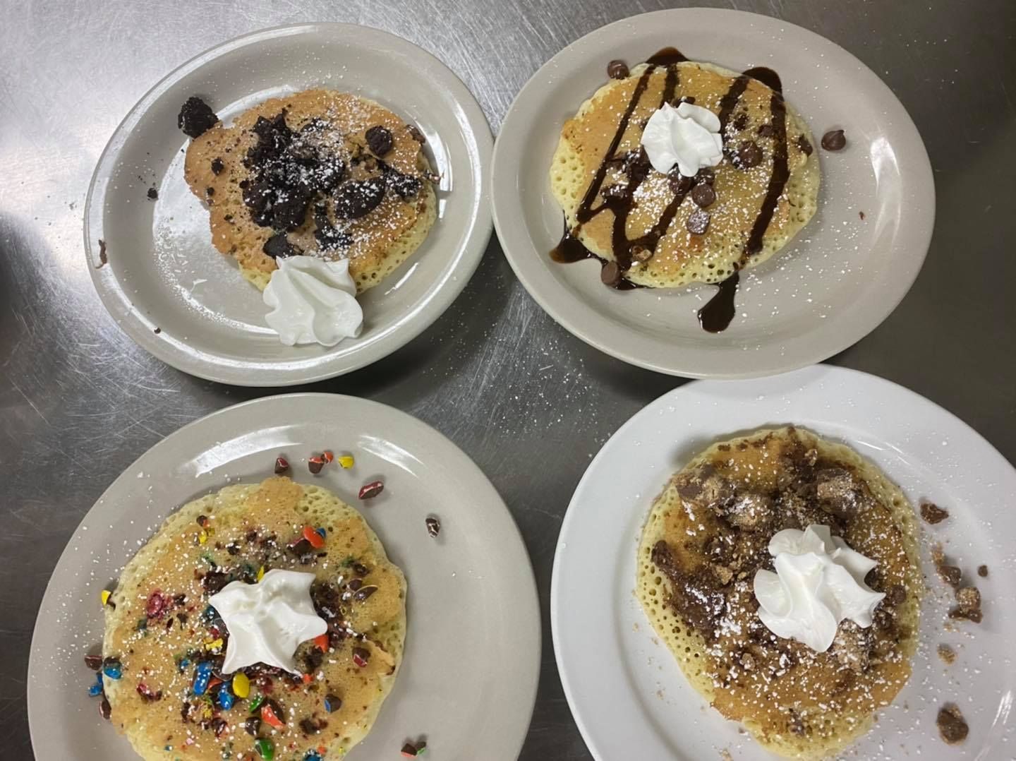 Four decorated pancakes on plates. Chocolate chips, candies, syrup, and whipped cream toppings.