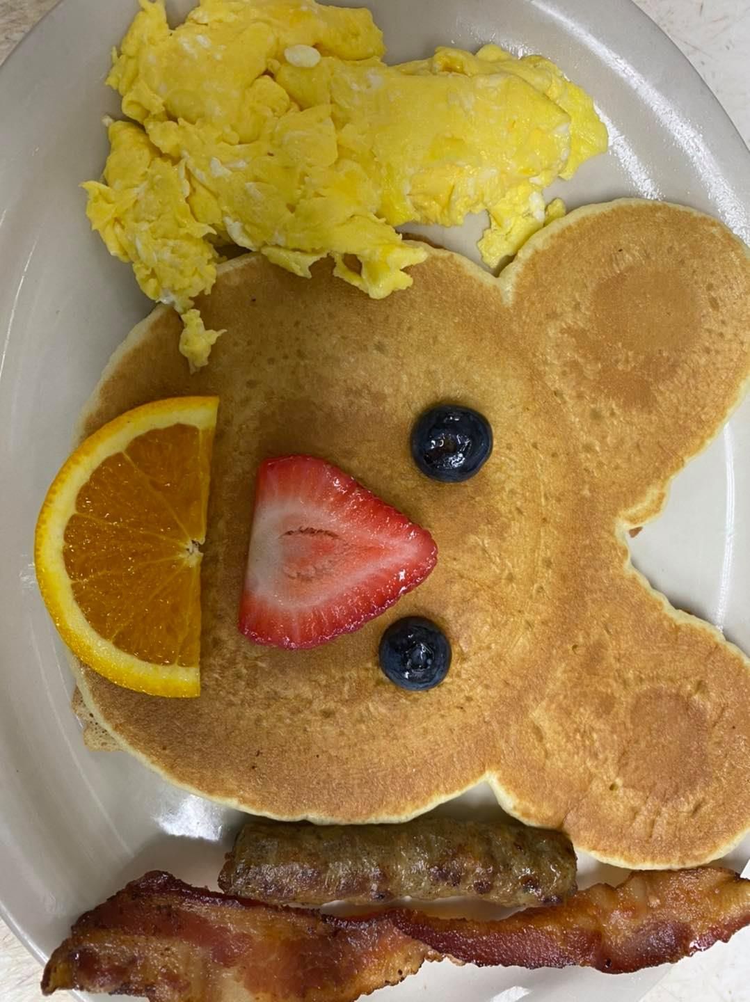 Bear-shaped pancake with fruit face, scrambled eggs, sausage, and bacon on a plate.