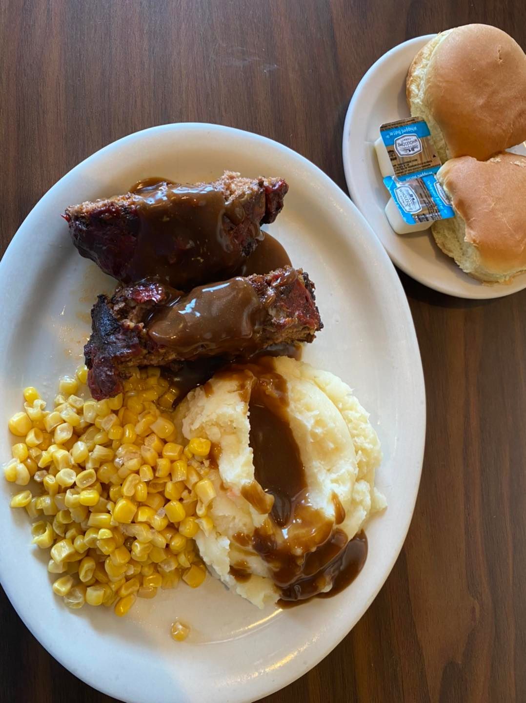 Meatloaf with gravy, mashed potatoes, corn, biscuits, and butter pats on a white plate.