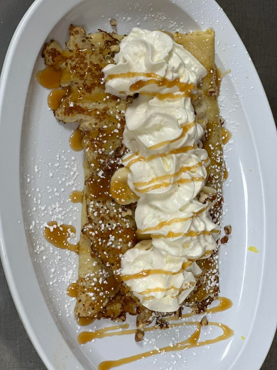 Crepe with whipped cream, caramel drizzle, bananas, and nuts on a white plate.