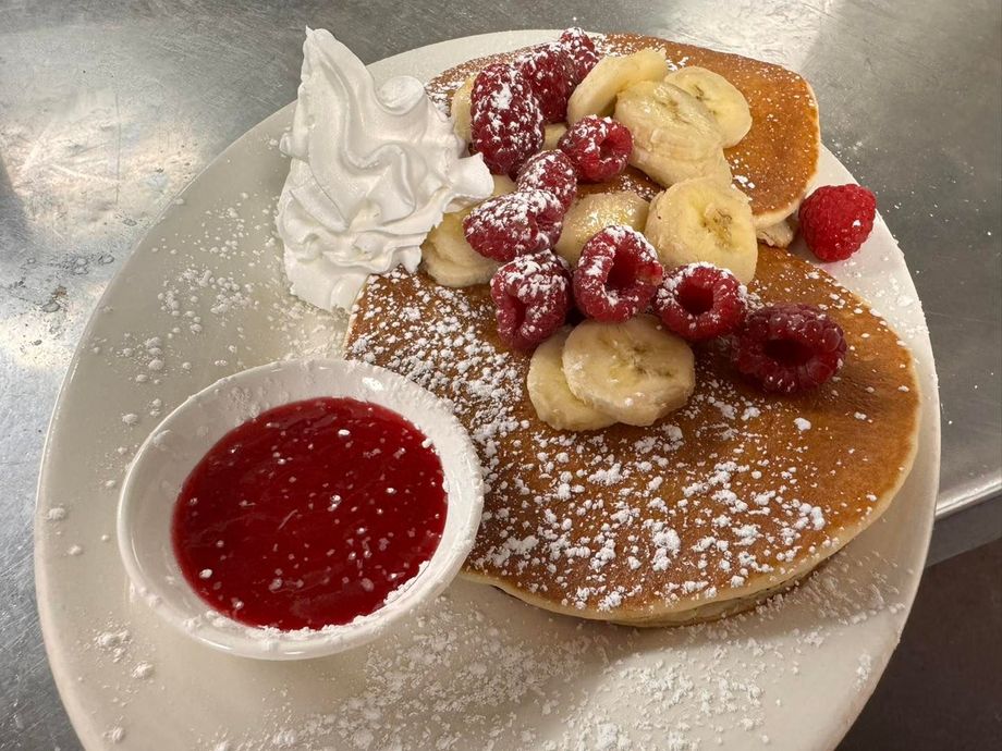 Pancakes topped with raspberries, banana slices, whipped cream, and raspberry sauce on a white plate.