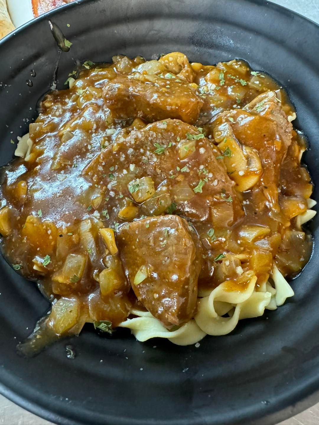 Beef and gravy served over noodles in a black bowl, with visible onions and mushrooms.
