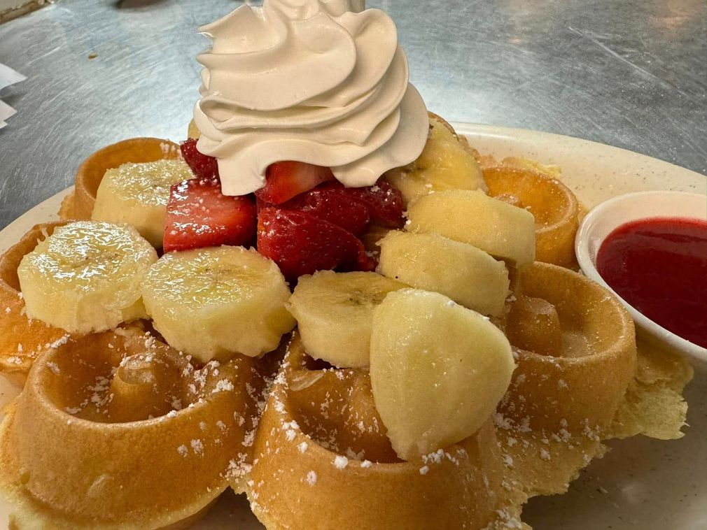 Waffle topped with whipped cream, strawberries, bananas, and powdered sugar. Served with a side of red syrup.