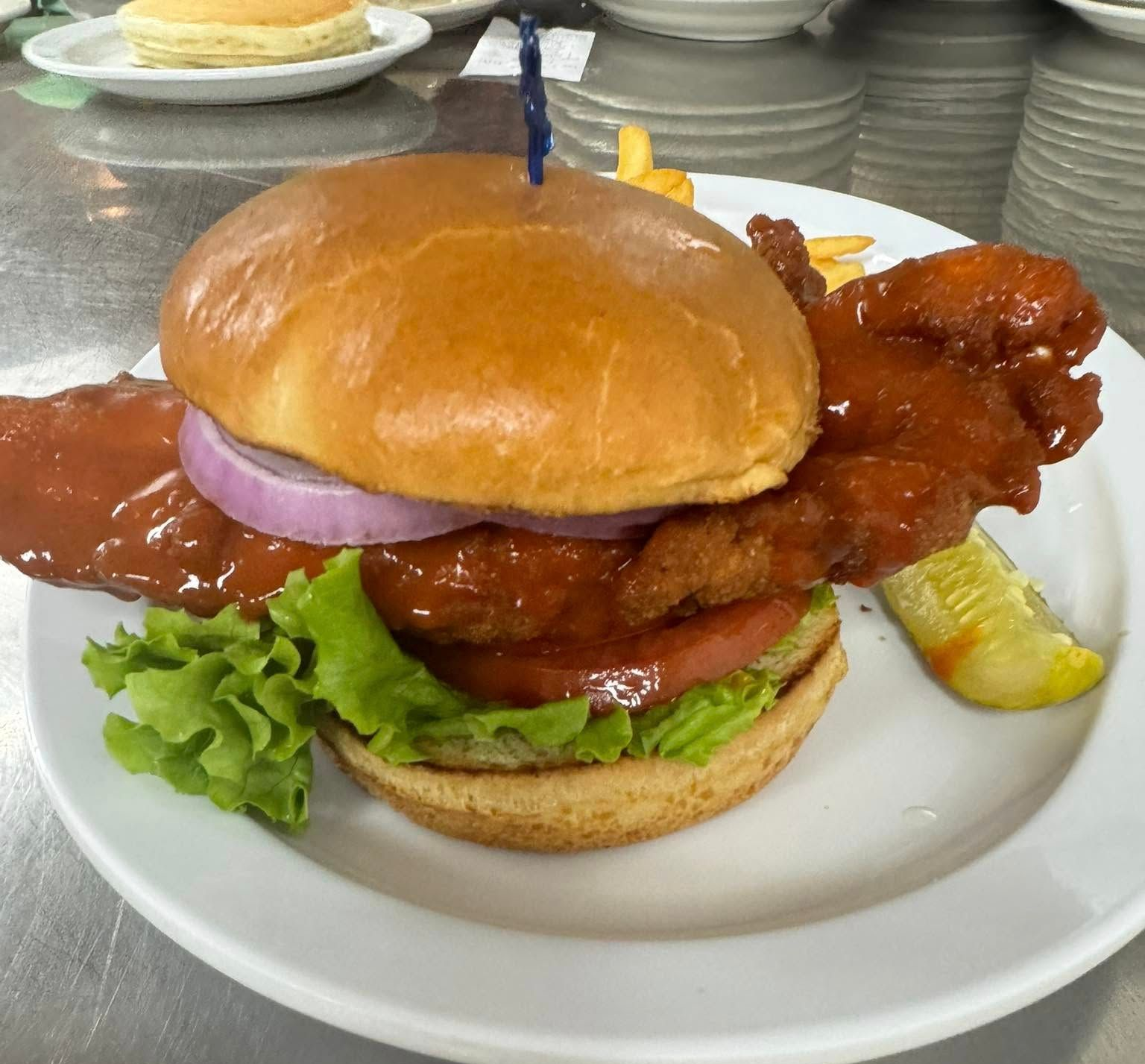 Spicy chicken sandwich with red sauce, lettuce, tomato, and onion on a plate with a pickle and fries.