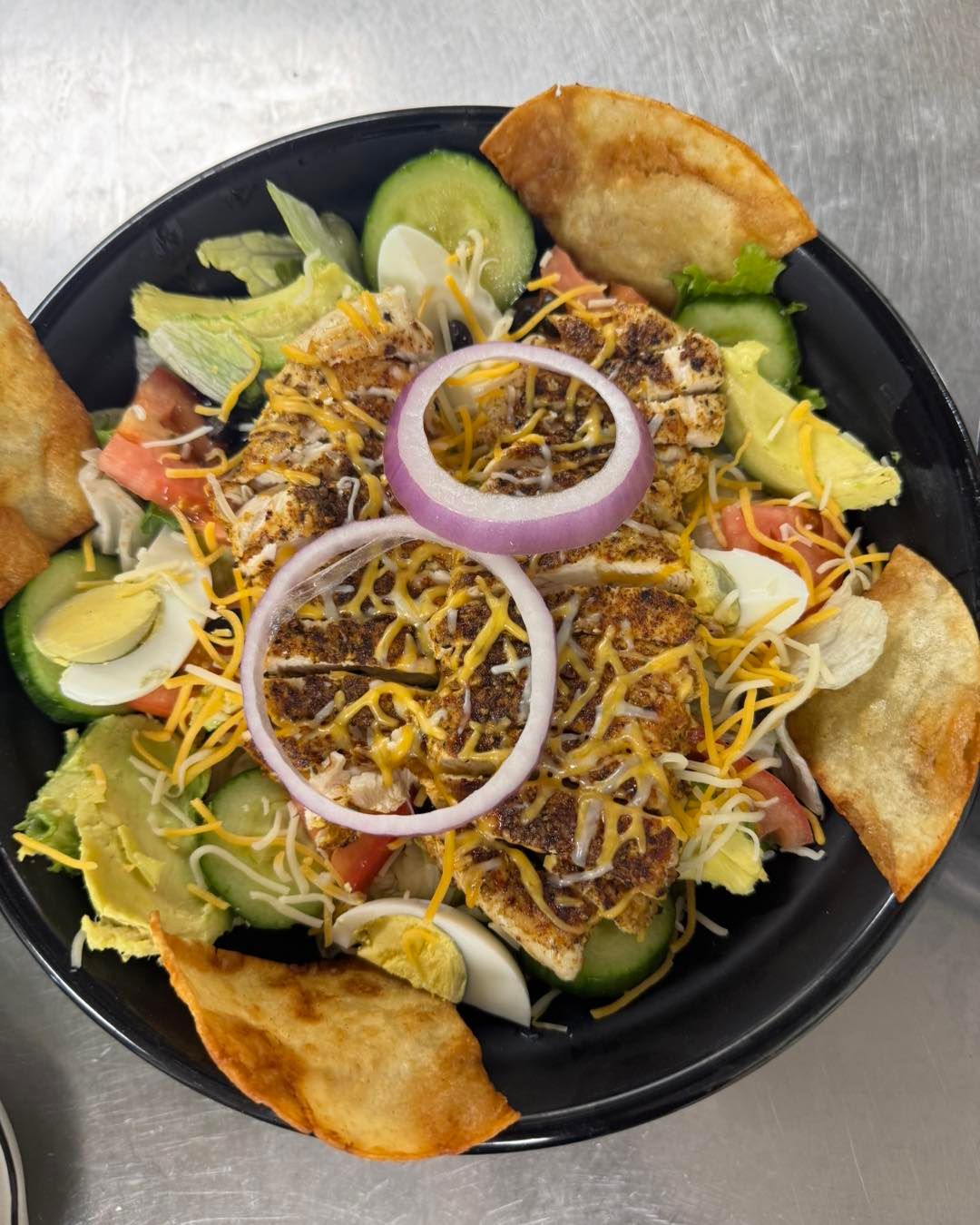 Chicken salad with eggs, tomatoes, cucumbers, cheese, red onion, and tortillas in a black bowl.