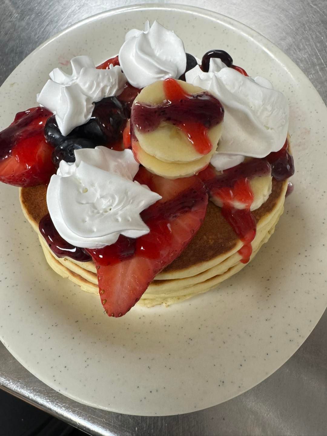 Stack of pancakes with fruit, whipped cream, and syrup on a plate.