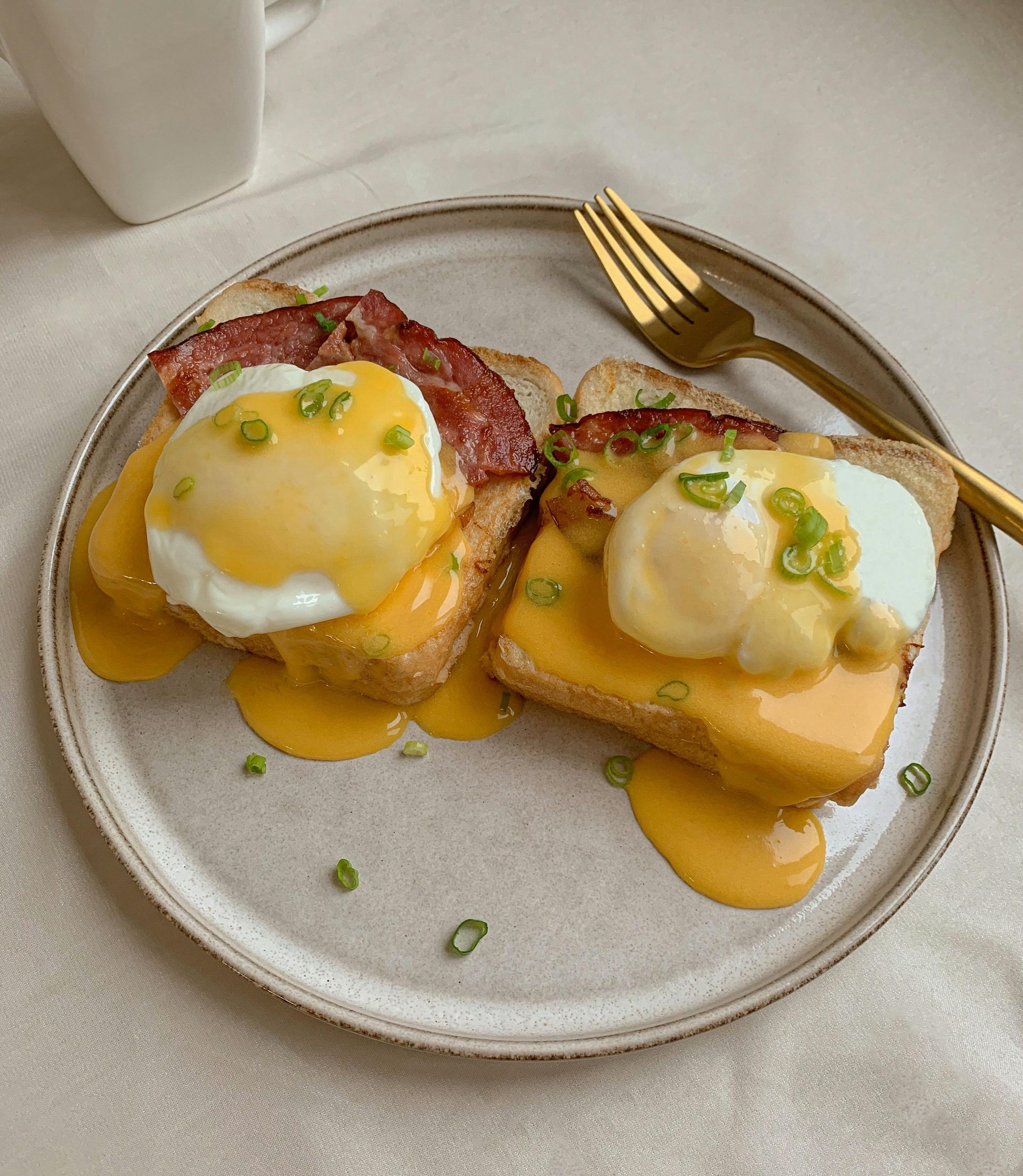 Eggs Benedict on toast, topped with hollandaise and bacon, on a plate with a gold fork.