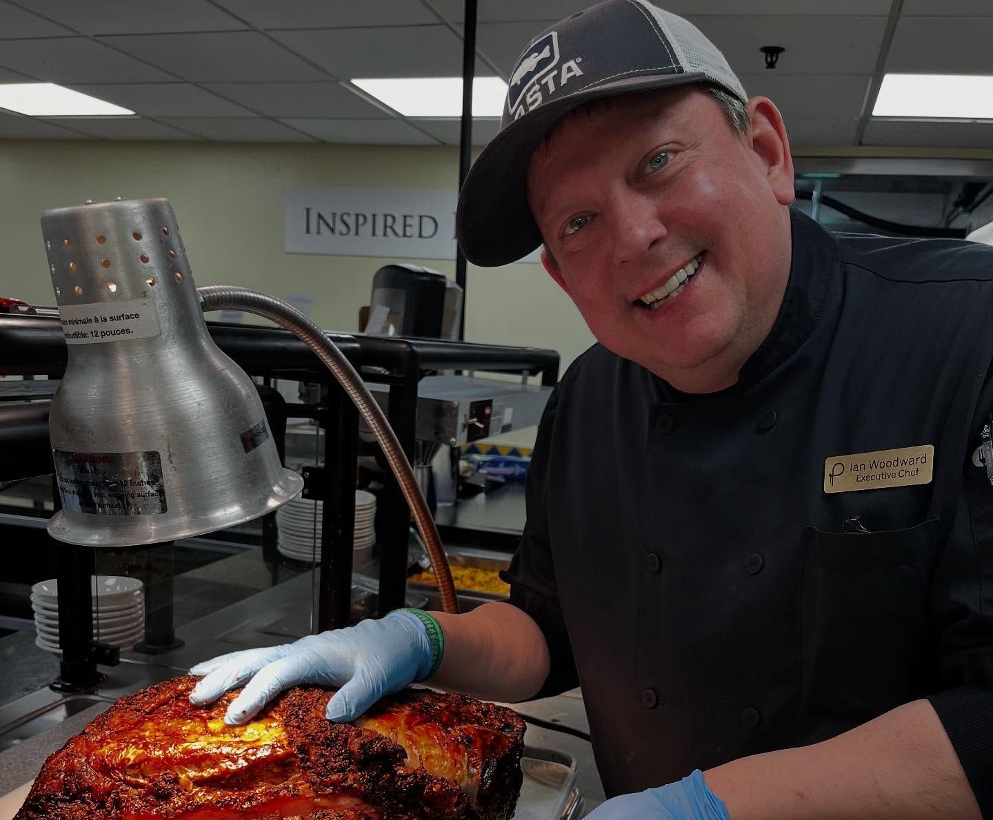Chef smiling, carving a roasted ham in a brightly lit kitchen, wearing a cap and gloves.